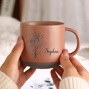 Può includere: Una tazza in ceramica marrone con un disegno floreale nero e il nome "Sophia" stampato sul lato. La tazza ha un fondo grigio.