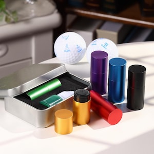 May include: An assortment of small, colorful metal containers and a silver tin with a black interior. The containers are in various colors, including gold, red, purple, blue, and black. Two golf balls are also present in the image.