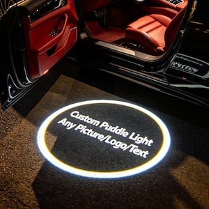 May include: Black luxury car with the door open, revealing a red interior. A custom puddle light illuminates the ground with the text "Custom Puddle Light Any Picture/Logo/Text" in a white font.