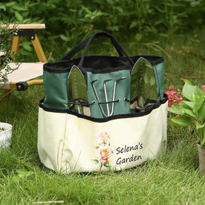 Pu&ograve; includere: Una borsa da giardinaggio verde e crema con manici neri, contenente attrezzi da giardinaggio. La borsa presenta un motivo floreale e il testo "Selena's Garden" stampato sul davanti. La borsa &egrave; sull'erba verde.