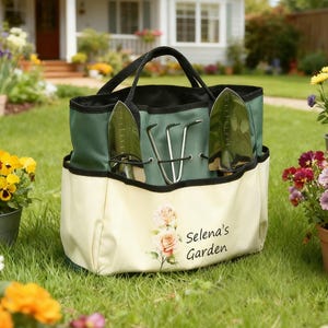 Pu&ograve; includere: Una borsa da giardinaggio verde e crema con manici neri, contenente due cazzuole e una forca a mano. La borsa presenta un motivo floreale e il testo "Selena's Garden". La borsa &egrave; su un prato.