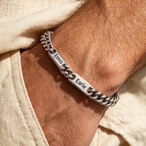 Könnte beinhalten: Ein silbernes Panzerkettenarmband mit zwei rechteckigen Namensschildern. Die Namensschilder sind mit den Namen "Ennis" und "Earle" graviert. Das Armband wird am Handgelenk getragen.