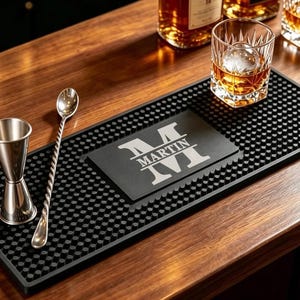 May include: A rectangular black rubber bar mat with a textured nub surface and a central rectangular inlay featuring the letter M and the name MARTIN in white text. A silver metal jigger and a long twisted bar spoon rest on the mat surface.