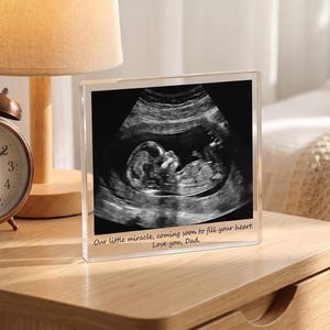 May include: A square clear acrylic block frame displaying a black and white ultrasound image of a fetus. The bottom of the frame features the printed text: Our little miracle, coming soon to fill your heart. Love you, Dad.