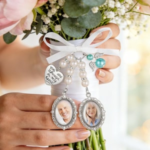 May include: A wedding bouquet charm featuring two oval silver-toned frames with portraits of an older man and woman. The charms hang from white pearl beads and a white ribbon, accompanied by a heart-shaped tag inscribed with you always in my heart.