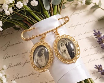Op de afbeelding: Een goudkleurige veiligheidsspeld-broche met twee ovale hangers met sierlijke geschulpte randen. Elke hanger bevat een zwart-witfoto van een persoon in trouwkleding, bevestigd aan een met wit lint omwikkeld handvat van een boeket.