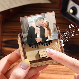 Puede incluir: Una caja de música de madera con una foto de una pareja abrazándose. La caja tiene un pequeño símbolo de nota musical y notas musicales dibujadas encima. La caja es sostenida por una persona con las uñas pintadas.