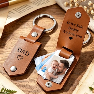May include: Two tan leather keychains with silver metal rings and snap buttons. One keychain is closed and engraved with DAD and two hearts. The other is open, revealing a photo of a person holding a baby and the text Drive Safe Daddy We love you.