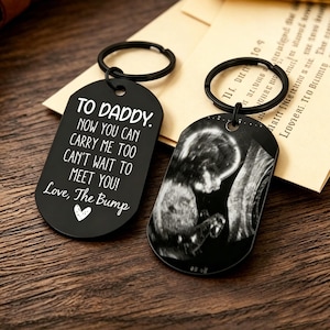 May include: Two black metal dog tag keychains with silver rings. One tag features white text reading: TO DADDY. NOW YOU CAN CARRY ME TOO CAN'T WAIT TO MEET YOU! Love, The Bump with a small heart. The other tag displays a black and white ultrasound image.