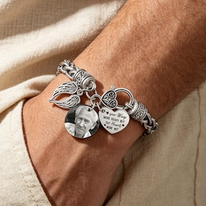 Peut inclure: Un bracelet en argent avec un design de chaîne. Le bracelet comprend un pendentif rond avec une photo en noir et blanc, un pendentif en forme de cœur avec du texte et un pendentif en forme d'aile. Le texte sur le pendentif en forme de cœur dit "Your Wings Were Ready But Our Hearts Were Not."