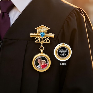 May include: A gold-colored graduation pin with a blue heart-shaped gem and the year 2026. The pin features a small photo pendant and the text "My Eternal Guardian Angel" on the back. The pin is attached to a black graduation gown.