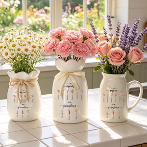 以下が含まれることがあります： 花で満たされた3つの白いセラミック花瓶。花瓶には「First Mom - Now Grandma」というテキストと名前が書かれています。花には、デイジー、ピンクのカーネーション、バラ、ラベンダーが含まれます。花瓶は白いタイルの表面にあります。