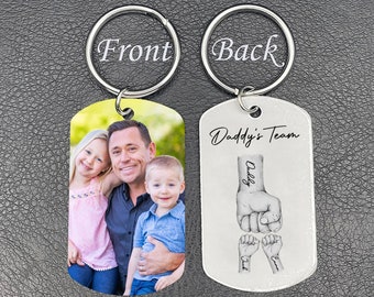 May include: A metal keyring featuring a photograph of a man and two children on the front. The reverse side of the keyring displays the text "Daddy's Team" and a fist illustration with the names "Daddy", "Dad", and "Emma".