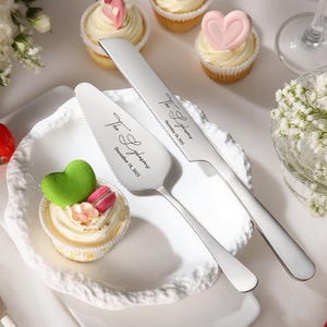 Może przedstawiać: Zestaw noża i serwera do ciasta ze stali nierdzewnej z grawerowanym tekstem. Zestaw jest prezentowany z babeczkami zwieńczonymi dekoracjami w kształcie serca. Tekst na nożu i serwerze brzmi "The [Last Name]" i "December 16, 2023".