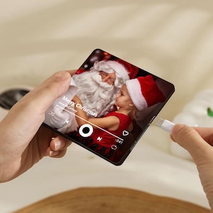 K&ouml;nnte beinhalten: Ein quadratischer digitaler Musikplayer mit einem Bildschirm, der ein Foto eines Kindes mit roter M&uuml;tze zeigt, das den Weihnachtsmann umarmt. Der Bildschirm zeigt den Text Merry Christmas, Jesse McCartney und Wiedergabesteuerungen. Ein wei&szlig;es USB-C-Kabel ist an der Seite eingesteckt.