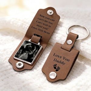 May include: A brown leather keychain with a silver key ring. The keychain features a rectangular pendant with a black and white ultrasound image. The text "Love You Daddy" and baby footprints are on the keychain. The case reads "Now you can carry me too. Can't wait to meet you! Love, little Bean."