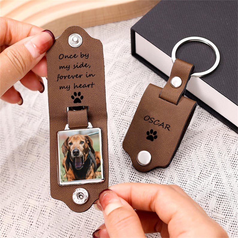May include: A brown leather keyring with a silver metal ring and a snap closure. The interior features a square photo of a brown dog and the text "Once by my side, forever in my heart" with a paw print. The exterior is engraved with "OSCAR" and a paw print.