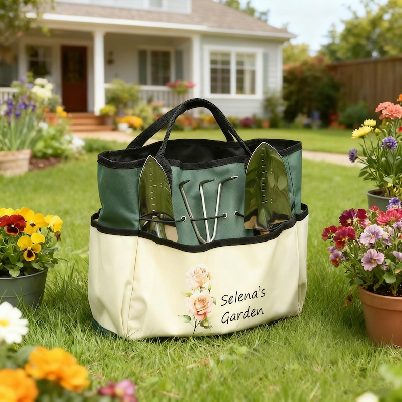 Pu&ograve; includere: Una borsa da giardinaggio verde e crema con manici neri, contenente due cazzuole e una forca a mano. La borsa presenta un motivo floreale e il testo "Selena's Garden". La borsa &egrave; su un prato.