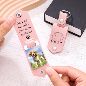 May include: A light pink leather keyring featuring a metal snap closure and a photo of a beagle. The interior is engraved with the text Once by my side, forever in my heart and a paw print. A second closed keyring shows a dog outline and the name OSCAR.
