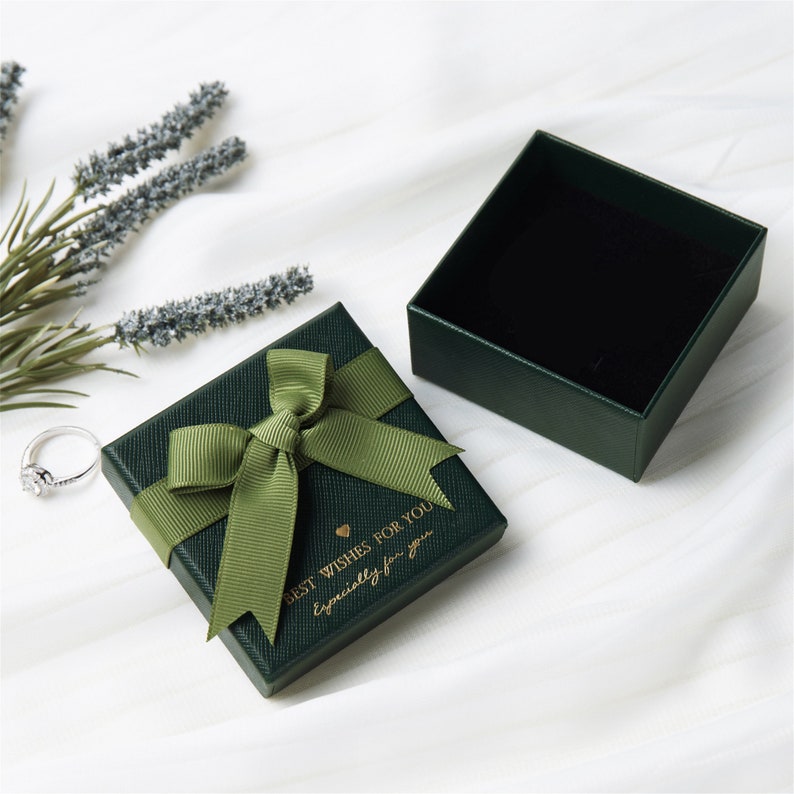May include: A square dark green jewelry gift box with a textured finish, topped with an olive green ribbon bow. The lid features gold text reading BEST WISHES FOR YOU and Especially for you. The box is open, revealing a black interior lining.