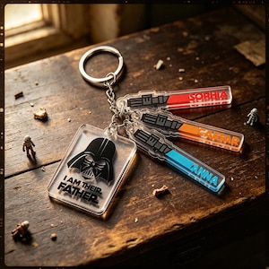 May include: A clear acrylic keyring featuring a black Darth Vader helmet graphic with the text I AM THEIR FATHER. Attached are three smaller rectangular acrylic charms shaped like lightsabers in red, orange, and blue, personalised with the names SOPHIA, CAESAR, and ANNA.