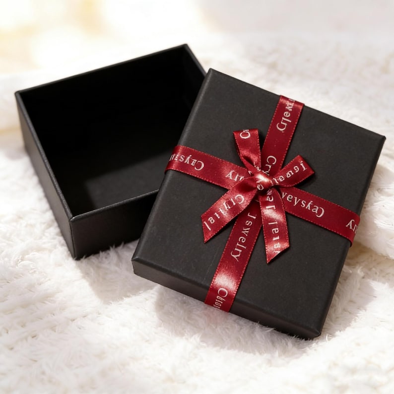 May include: A square black gift box with a matching lid placed beside it. The lid is adorned with a dark red satin ribbon tied in a bow, featuring the white text "Cysy Jewelry" printed repeatedly along the length of the ribbon fabric.