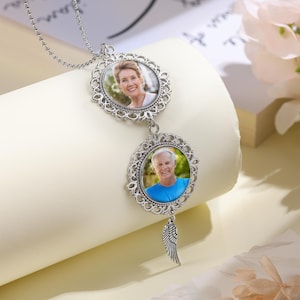 May include: A silver-colored, double-sided photo pendant necklace. The pendant features two oval frames with ornate filigree edges, each holding a color photograph. A small wing charm hangs below the bottom frame. The necklace chain is a delicate ball chain.