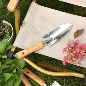 Pu&ograve; includere: Una cazzuola da giardino in acciaio inossidabile con manico in legno, incisa con "Lily's Garden". Poggia su una sedia beige, circondata da fiori rosa e fogliame verde. Uno strumento da giardinaggio per piantare e trapiantare.