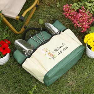 Pu&ograve; includere: Una borsa da giardinaggio verde e crema con attrezzi da giardinaggio. La borsa ha un manico nero e il testo "Selena's Garden" con un motivo floreale. Gli attrezzi includono una cazzuola e un rastrello a mano. La borsa &egrave; sull'erba.