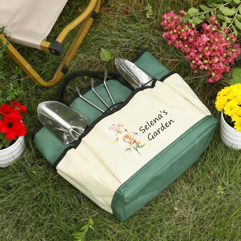 Pu&ograve; includere: Una borsa da giardinaggio verde e crema con attrezzi da giardinaggio. La borsa ha un manico nero e il testo "Selena's Garden" con un motivo floreale. Gli attrezzi includono una cazzuola e un rastrello a mano. La borsa &egrave; sull'erba.