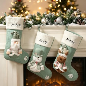 Pode incluir: Três meias de Natal verde-sálvia com punhos de pele sintética branca, personalizadas com os nomes Jackson, Avery e Millie. As meias apresentam aplicações de pelúcia de um gnomo, um boneco de neve e uma rena, com flocos de neve brancos.