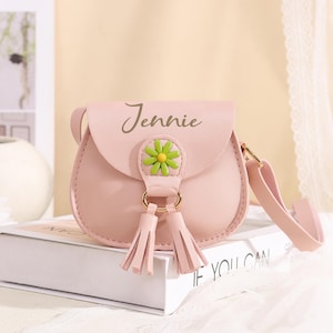Può includere: Una borsa a tracolla rosa chiaro con il nome Jennie stampato in corsivo scuro sulla patta anteriore. La borsa è decorata con un ornamento a forma di margherita verde e gialla e due nappe rosa abbinate appese a un anello in metallo dorato.