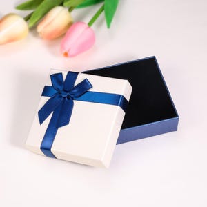 Puede incluir: Una caja de regalo cuadrada para joyas con una tapa de color crema que presenta un lazo de sat&eacute;n azul oscuro. La base de la caja es de color azul oscuro con un forro interior negro. La caja se muestra abierta con la tapa al lado.