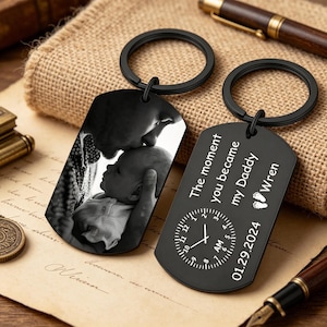 Può includere: Due portachiavi neri a forma di targhetta per cani. Uno presenta una foto in bianco e nero di un genitore che bacia un bambino. L'altra targhetta riporta la scritta "The moment you became my Daddy Wren" con un design a orologio e la data 01.29.2024.
