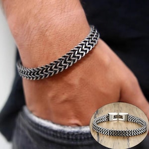 May include: A silver bracelet with a woven chain pattern is shown on a wrist. The bracelet features a secure clasp. A smaller image shows the bracelet with the names "Dad" and "Anna" engraved on the clasp.