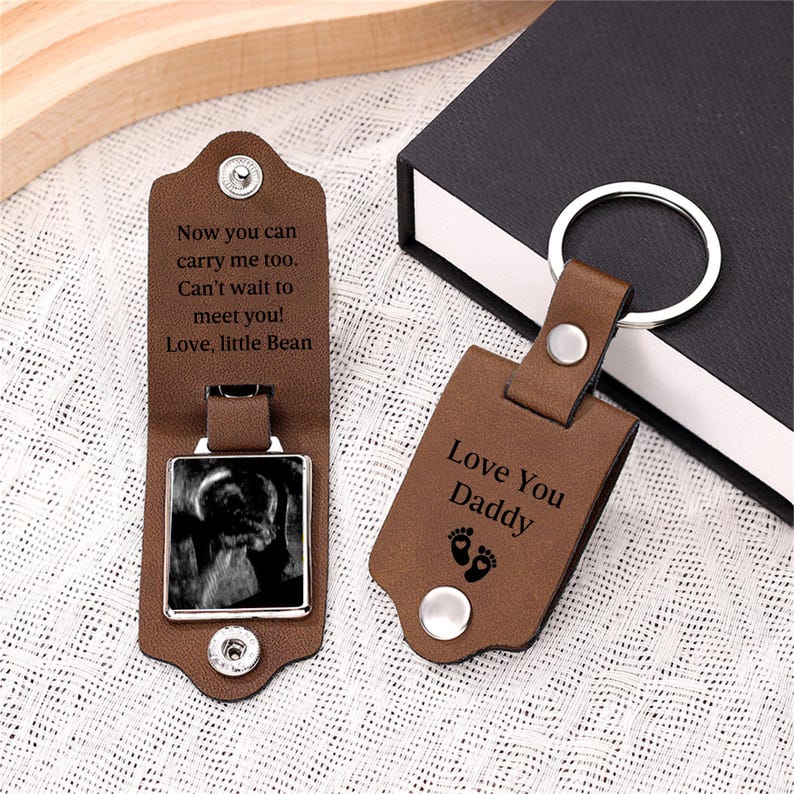 May include: Two brown leather keychains with silver metal rings and snap closures. One keychain is open, revealing a black and white ultrasound image and the text: Now you can carry me too. Can't wait to meet you! Love, little Bean. The other is closed, displaying: Love You Daddy with two small footprints.
