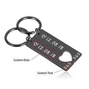 May include: Black metal keychain with two interlocking key rings. The keychain has two rectangular tags with the text "&hearts; 12.08.18" and "B&G" engraved on them.