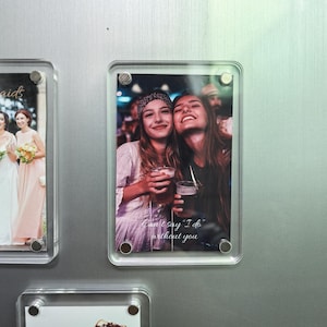 Puede incluir: Un marco de fotos magn&eacute;tico rectangular de acr&iacute;lico transparente que contiene una fotograf&iacute;a de dos personas sonrientes sosteniendo vasos, con el texto blanco "Can't say "I do" without you" impreso en la parte inferior. El marco est&aacute; asegurado por cuatro pernos de esquina de metal plateado.