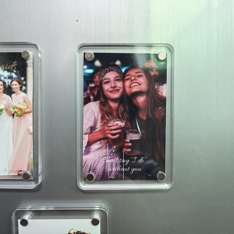 Puede incluir: Un marco de fotos magn&eacute;tico rectangular de acr&iacute;lico transparente que contiene una fotograf&iacute;a de dos personas sonrientes sosteniendo vasos, con el texto blanco "Can't say "I do" without you" impreso en la parte inferior. El marco est&aacute; asegurado por cuatro pernos de esquina de metal plateado.