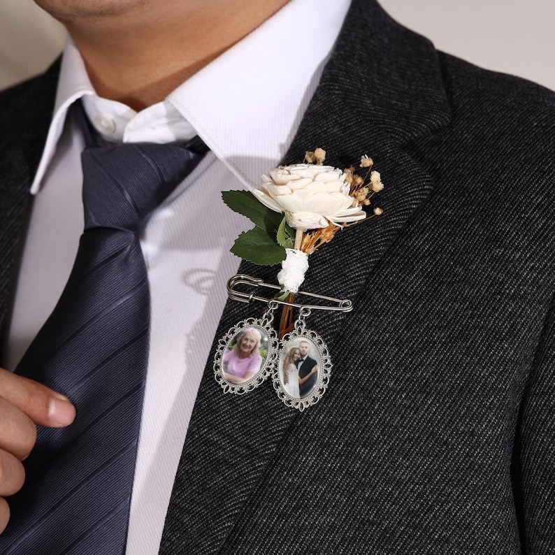 May include: A boutonniere featuring a white flower, green leaves, and dried accents attached to a silver safety pin. Two oval-shaped silver frames with intricate filigree edges hang from the pin, each containing a small photograph of a person.
