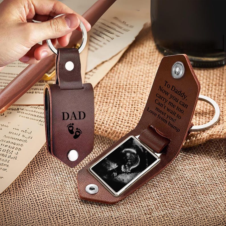 May include: A brown leather keychain with a metal ring, featuring the word DAD and baby footprints on the front. The interior reveals a rectangular frame holding a black and white ultrasound image and text that reads To Daddy, Now you can carry me too. Can't wait to meet you! Love from bump.
