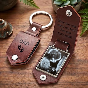 Può includere: Un portachiavi in pelle marrone con un anello in metallo argentato. Il lato chiuso presenta la scritta DAD e due impronte di piedi di neonato. Il lato aperto rivela una cornice rettangolare con un'ecografia in bianco e nero e il testo Now you can carry me too! See you very soon!
