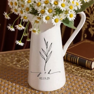 May include: White ceramic pitcher with a handle, filled with daisies. The pitcher has a black leaf design and the initials "j + h" with the date "02.15.25" in black script. The pitcher is on a woven surface.
