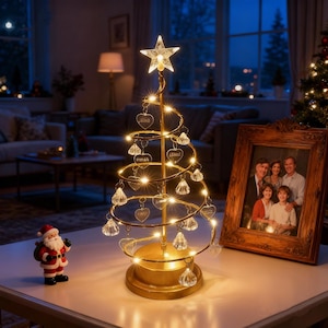 Puede incluir: Un árbol de Navidad en espiral de color dorado con una estrella en la parte superior y adornos en forma de corazón. El árbol está iluminado con luces blancas cálidas y se encuentra sobre una mesa junto a una foto familiar enmarcada y una figura de Papá Noel.