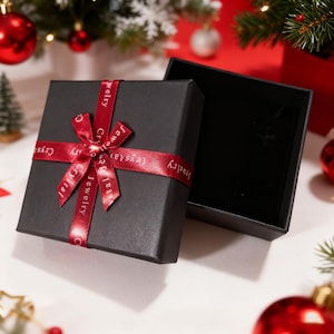 May include: A square black jewellery gift box with a removable lid. The lid is decorated with a red satin ribbon tied in a bow, featuring the repeating white text Jewelry Crystal. The interior of the box is lined with black velvet material.