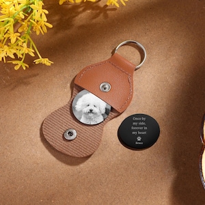 May include: A brown leather keychain with a silver ring, holding a round photo of a fluffy white dog. A black, oval-shaped charm next to it reads "Once by my side, forever in my heart" with a paw print and the name "Bruce."