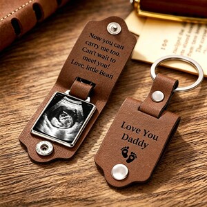 May include: A brown leather keychain with the text "Love You Daddy" and baby footprints. The keychain also features a small metal frame holding a sonogram image. The text "Now you can carry me too. Can't wait to meet you! Love, little Bean" is also visible.