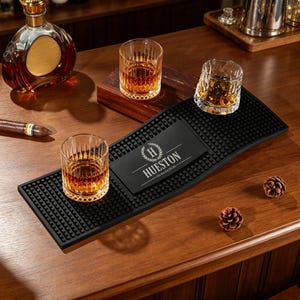 Könnte beinhalten: Eine rechteckige schwarze Gummimatte für die Bar mit strukturierter Noppenoberfläche und einem mittigen Feld, das den Text HUESTON mit einem Lorbeerkranz-Emblem zeigt. Zwei klare Gläser mit bernsteinfarbener Flüssigkeit stehen auf der Matte.