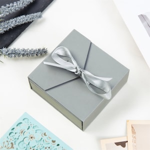 May include: A gray gift box with a silver ribbon tied in a bow. The box is made of cardboard and has a smooth finish.