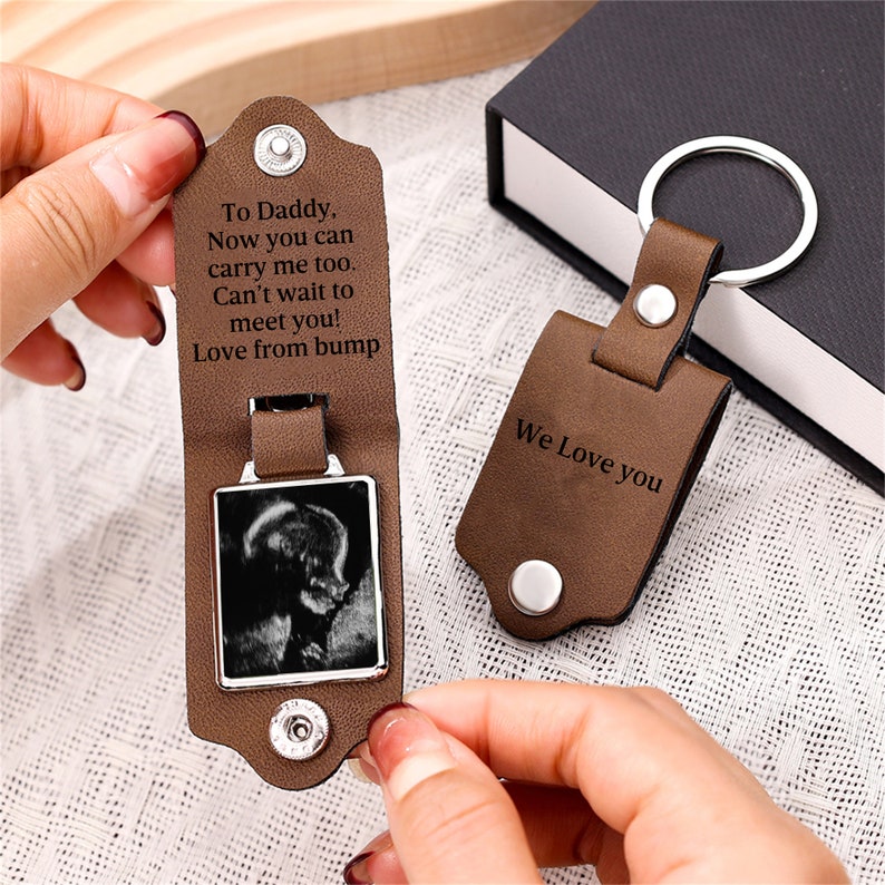 May include: A brown leather keychain with a silver metal ring and snap closures. The interior features a black and white ultrasound image and the text: To Daddy, Now you can carry me too. Can't wait to meet you! Love from bump. The exterior says We Love you.